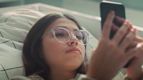 zoom de una adolescente caucásica navegando por el teléfono mientras está acostada en la cama de espaldas