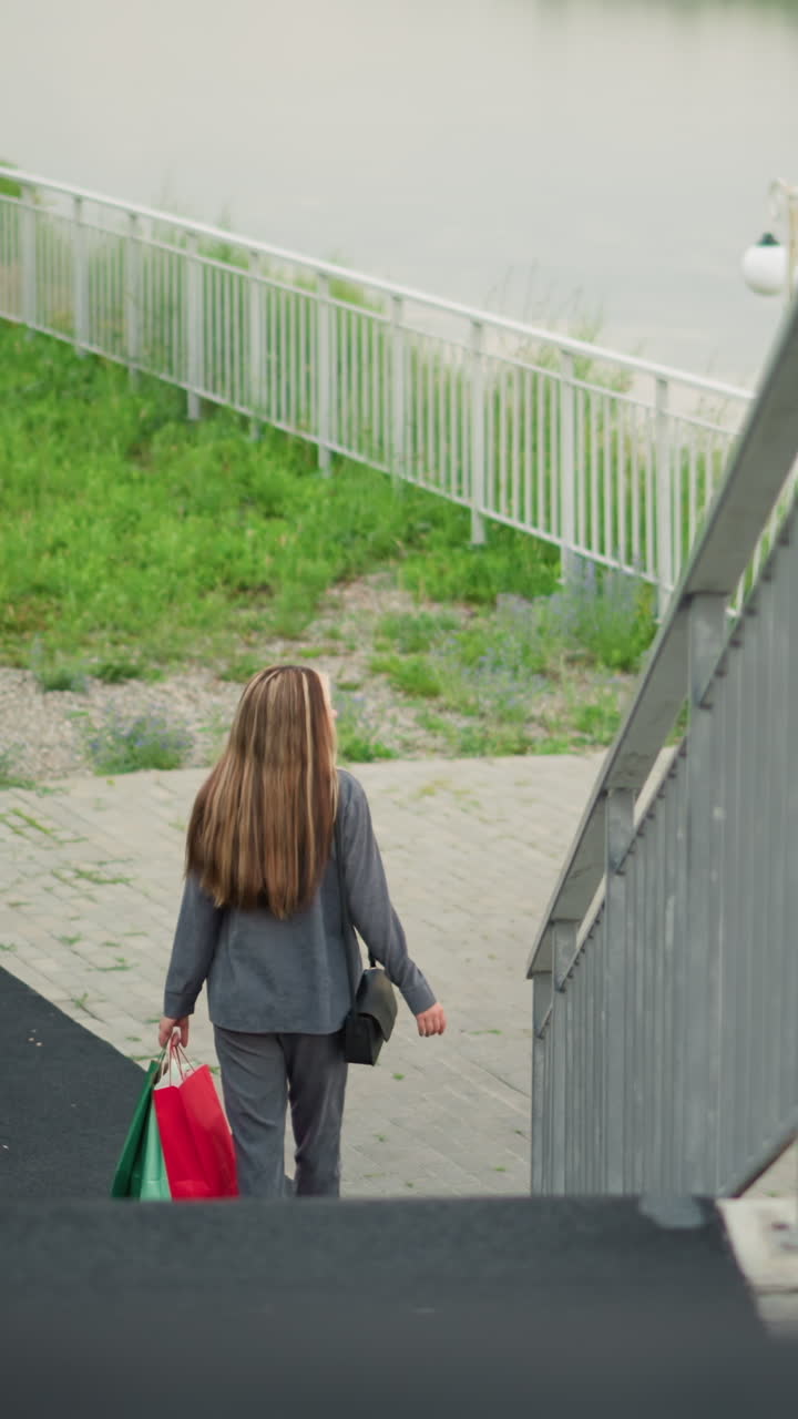 계단 아래로 내려가는 동안 다채로운 쇼핑 가방을 들고 계신 여인의 뒷면, 뒷면에는 강, 램프 기둥, 초록색을 특징으로 한 세 명의 개인이 서 있습니다.