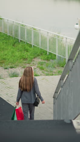 vista trasera de una mujer sosteniendo bolsas de compras coloridas mientras baja las escaleras, sosteniendo una barandilla para el apoyo, con tres personas en el fondo, con un río, un poste de lámpara y vegetación