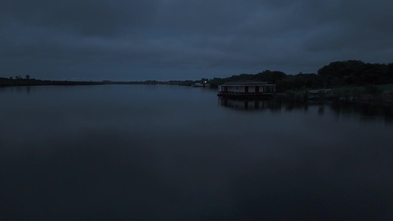 Atmospheric drone view over the Amazon River at night in South America, with calm dark water, a remote houseboat and tropical vegetation beneath a brooding sky