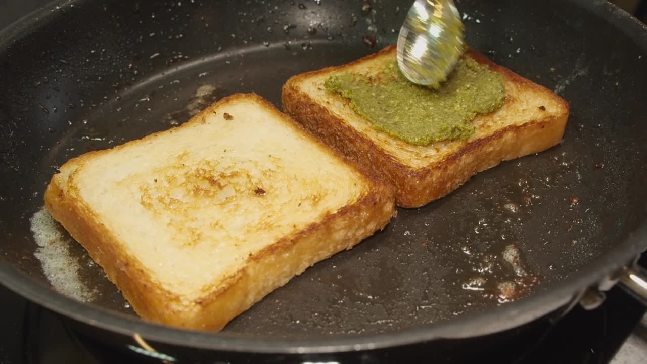 Green pesto is spread onto golden brown bread frying in hot skillet