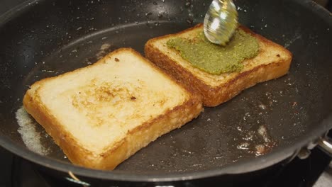 Green-pesto-is-spread-onto-golden-brown-bread-frying-in-hot-skillet