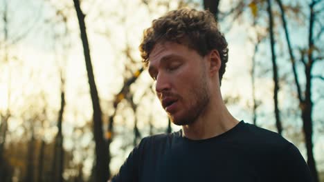 A-confident-and-serious-man-with-curly-hair-and-a-beard-in-a-black-sports-uniform-wipes-sweat-from-his-forehead-with-a-gray-towel-after-an-evening-jog-in-the-autumn-forest