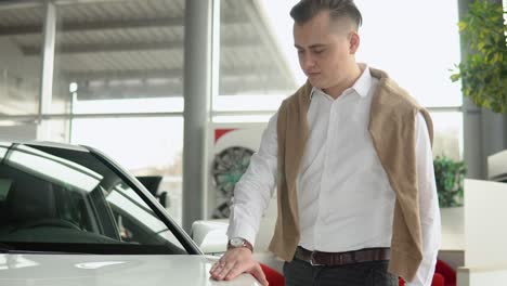 guy touches the hood of a shiny white car. young man looking carefully at car in car dealership