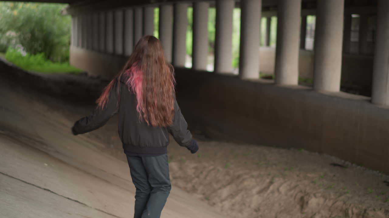 Ragazza giocosa che attraversa un passaggio colonnato. Foto spontanea di una donna con ciocche rosa che cammina in un corridoio. Giovane donna con vivaci mèches rosa che esplora naturalmente un passaggio sotterraneo.