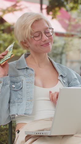 happy rich young woman using laptop winner victory holding cash money in euro banknotes in city