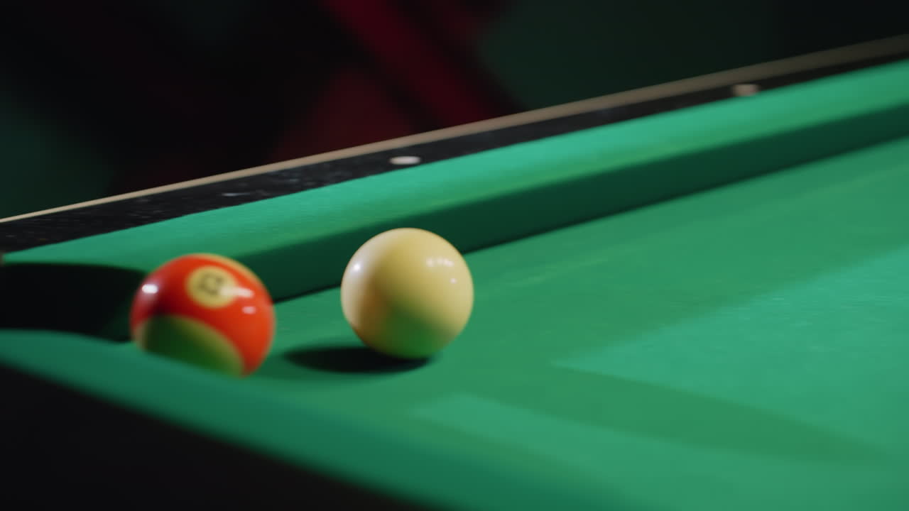 Cue stick strikes white billiard ball on green pool table. Other colorful billiard balls scattered across table. Captures precision and movement in strategic billiards play with sharp focus on cue