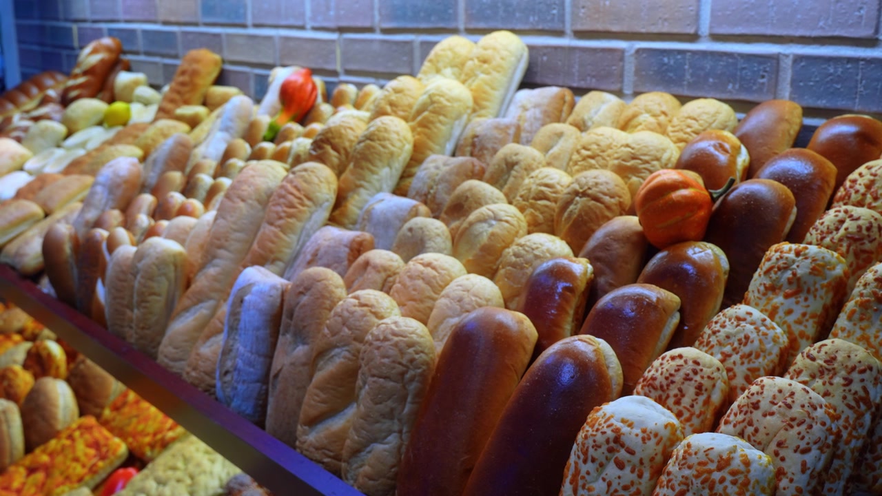 Close up view of assorted freshly baked bread rolls on a bakery shelf. A variety of golden buns, shiny brioche, and cheese topped loaves are arranged neatly with a small decorative pumpkin.