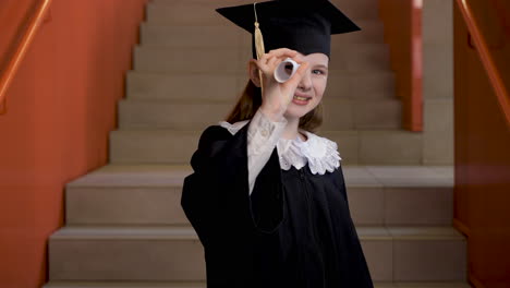 Happy-Preschool-Female-Student-In-Cap-And-Gown-Dancing-And-Playing-With-Graduation-Diploma-While-Looking-At-The-Camera