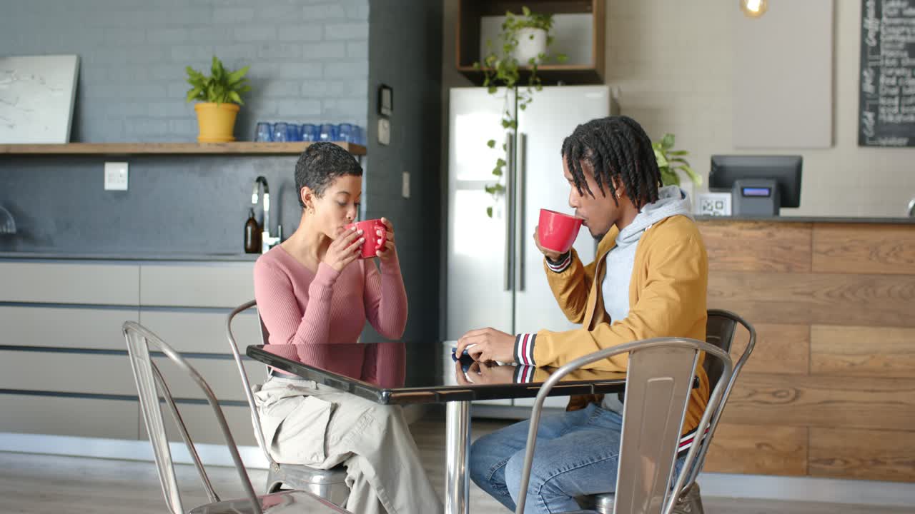 Diverse couple sipping from red mugs and chatting at cafe table, waitress crossing behind counter