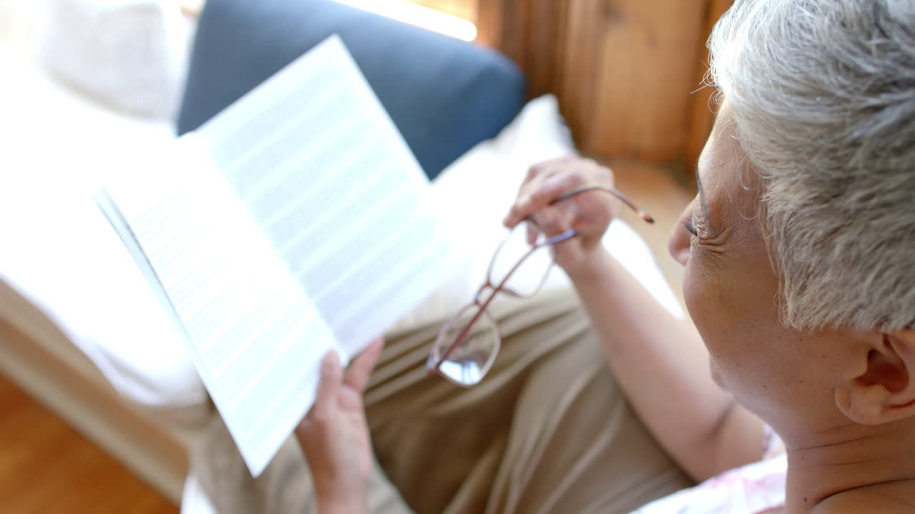 una mujer bi-racial feliz en el sofá leyendo un libro en la ventana de su casa, en cámara lenta.
