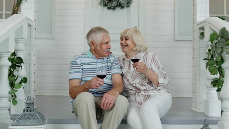 una pareja de ancianos caucásicos bebiendo vino en el porche de su casa, mirando a la cámara y sonriendo.