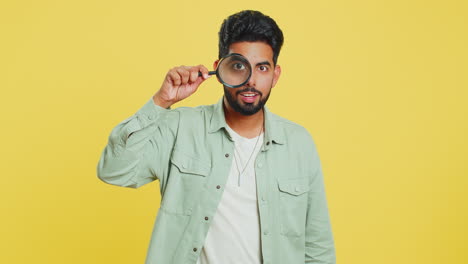 indian young man holds magnifying glass near face looking at camera with big zoomed eye, analyzing