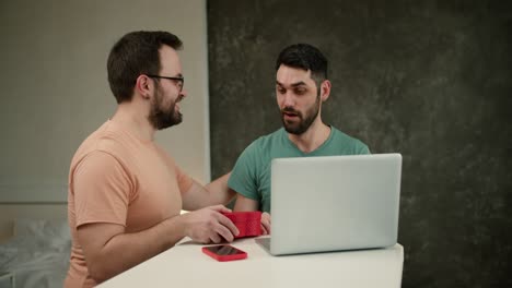 Handsome-young-man-using-laptop-and-smiling-while-boyfriend-holding-gift-box-for-him