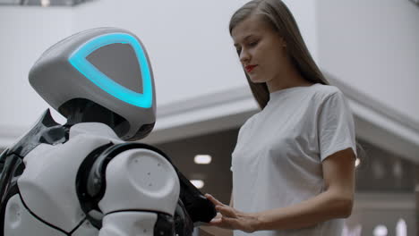 Smiling-girl-in-white-t-shirt-is-in-contact-with-humanoid.-Cyborg-helps-the-girl-answering-questions.-Useful-robot-assistant-in-the-office-and-shopping-center