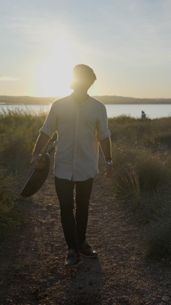 A Man with a Guitar Walking by a Lake at Sunset