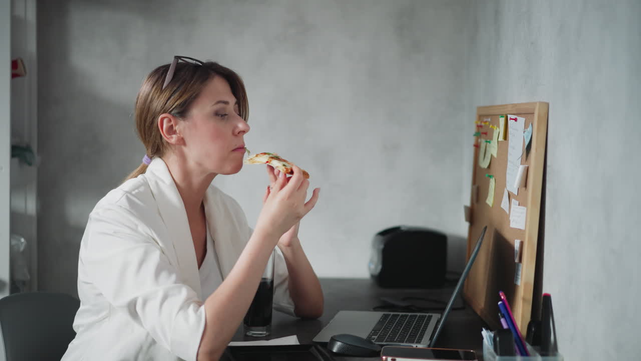 Remote worker with hair tied back and eyeglasses on head enjoys slice of pizza while working at laptop in home office space with cork board full of sticky notes in background