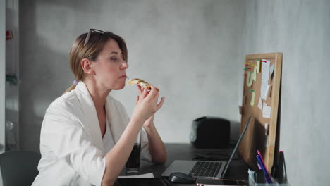 remote worker with hair tied back and eyeglasses on head enjoys slice of pizza while working at laptop in home office space with cork board full of sticky notes in background