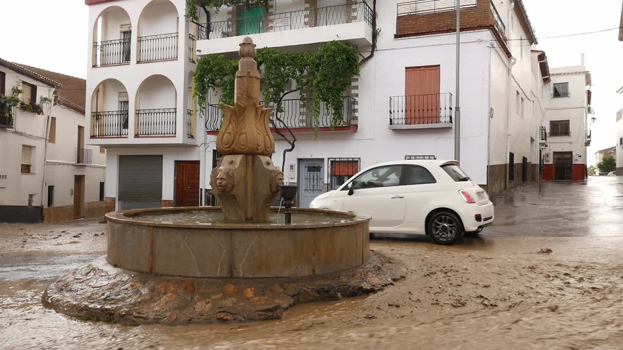 Water and mud fill a street during a severe flood in a Spanish village. A white car struggles to navigate past an ornamental stone fountain