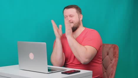 joven hombre de negocios feliz en una camiseta rosa trabaja en una computadora portátil y aplaude sus manos en una pantalla verde. retrato de un hombre masculino hablando mirando a su computadora portable. hombre trabajando en su escritorio en la oficina