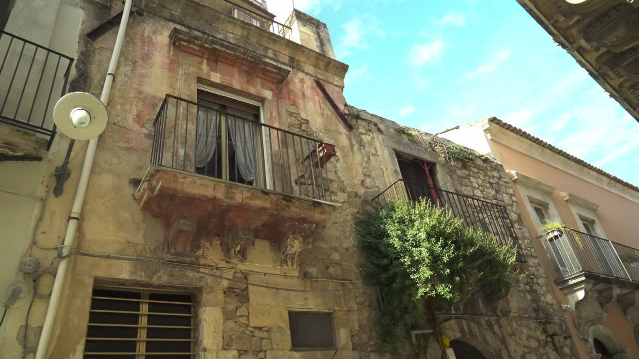Vista de rua em ângulo baixo de edifícios de pedra envelhecidos e varandas com grades de ferro ornamentadas em Ragusa, Sicília, Itália, arquitetura barroca típica sob um céu azul.