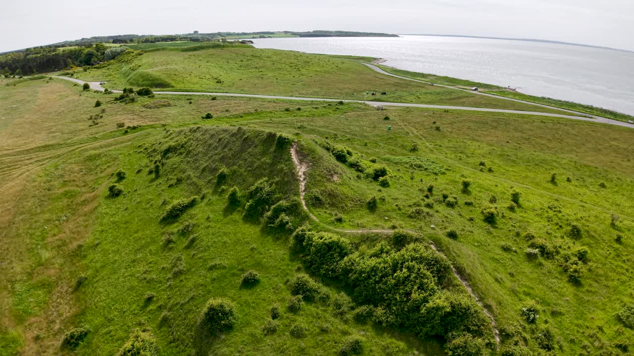 Aerial drone footage of Denmark’s coastal landscape with rolling green hills, grassy terrain, and calm shoreline under cloudy daylight