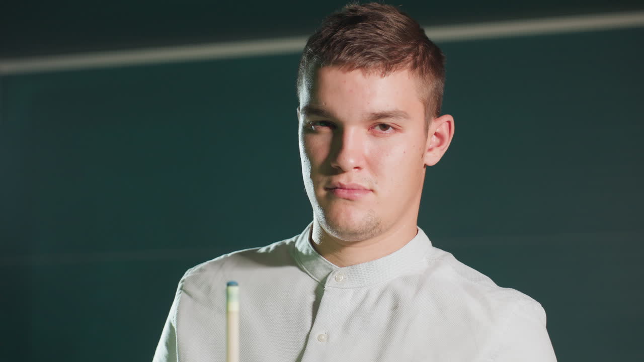 крупный план игрока в белой рубашке, держащего клюшку, нанося красную обложку с сосредоточенным выражением. темный фон усиливает драматическое освещение, подчеркивая концентрацию и подготовку к игре