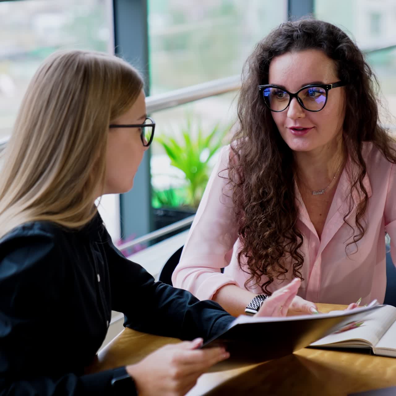 Brunette and blonde long-haired ladies wearing glasses sit at table talking. Female colleagues discussing job moments