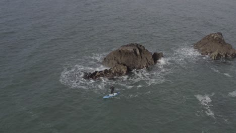 aerial of sup athlete paddling in chaotic water near rocky ocean islet