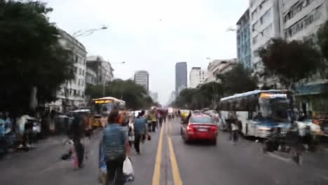 a lively urban street is bustling with pedestrians and vehicles under gray skies. rain falls as people navigate through the crowds, surrounded by tall buildings and vibrant activity.