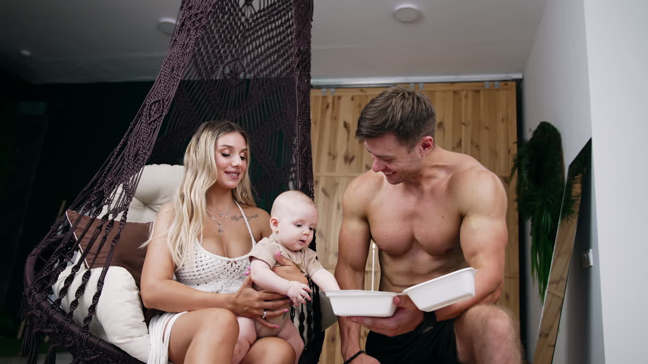 Young Caucasian family with a newborn celebrating holiday. Dad lights the candle on the cake and baby sitting on mom's laps leans to the candle.