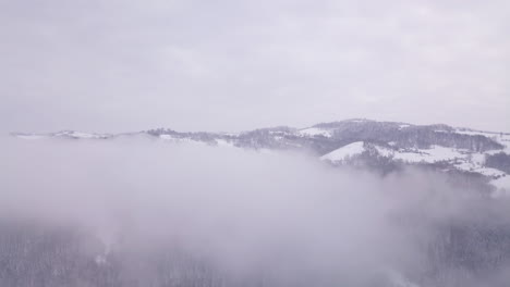 Aerial-footage-above-clouds-showing-a-hill-covered-by-snow-at-the-end-of-them-and-some-trees-covered-by-snow-under-the-clouds