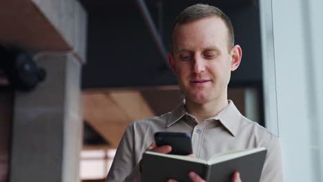 mature man holds the notebook opened in front of him and print something on the phone. he throws interested look into the window.