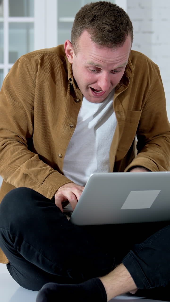 Young man looks mad working long hours online. Guy sitting on a kitchen table and typing on a wireless laptop. Funny fellow playing computer games at home. Vertical video