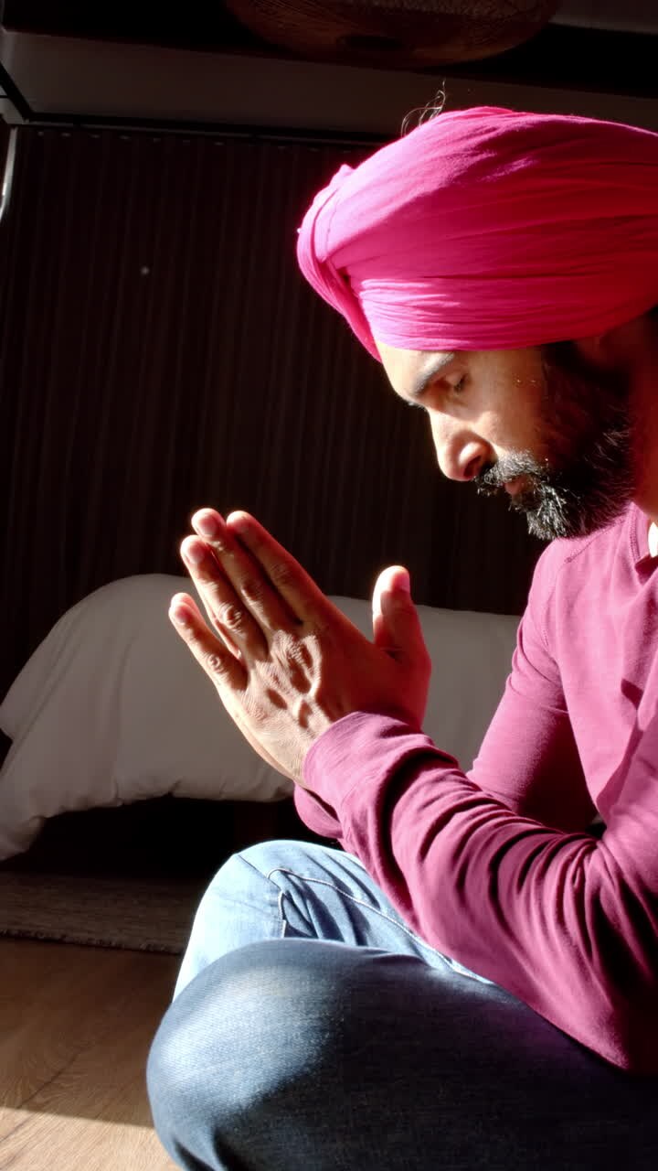 Vertical video: Praying with hands together, man in turban in traditional attire sitting at home