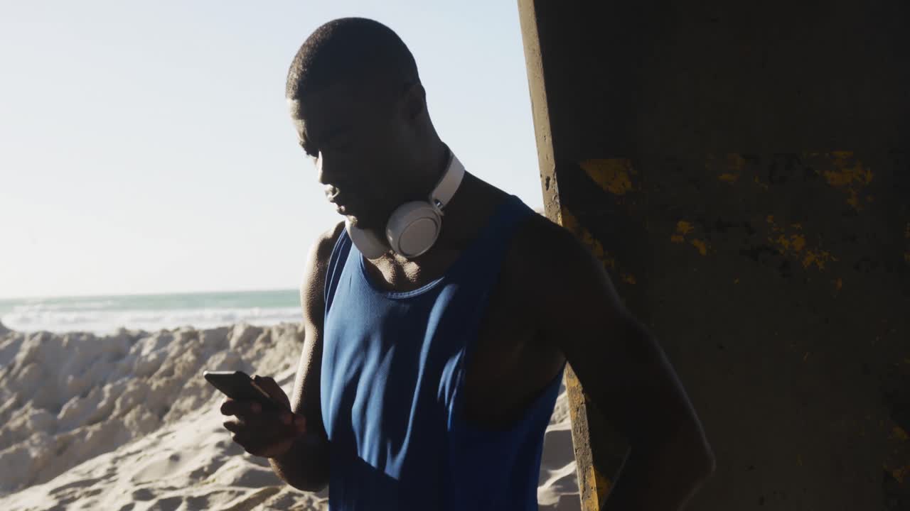 Afrikansk-amerikansk mand tjekker sin smartphone og tager en pause under træning udendørs ved havet.