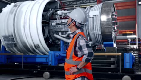 side view of asian male engineer wearing safety helmet looking around while standing with arms akimbo with airplane engine maintenance conducted, analytics checking the turbine
