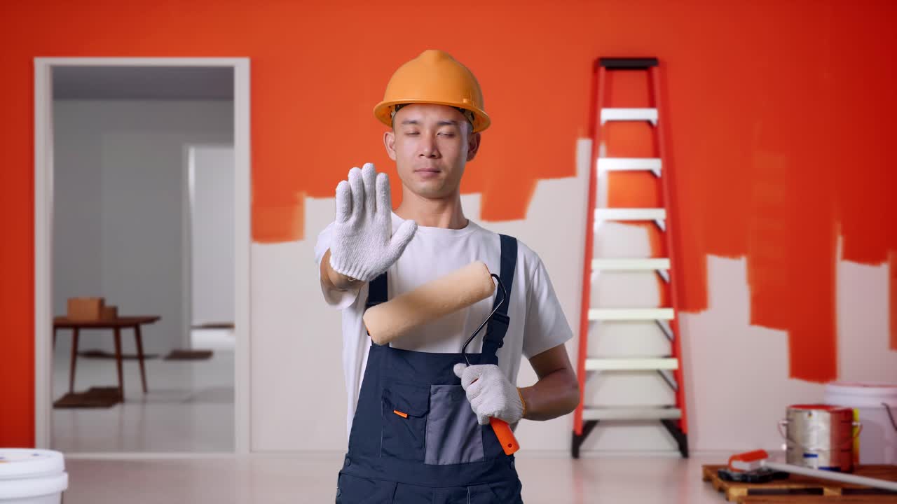 pintor hombre asiático con casco de seguridad mirando a la cámara y desaprobando sin señal de mano mientras está de pie con la pared de pintura en la habitación