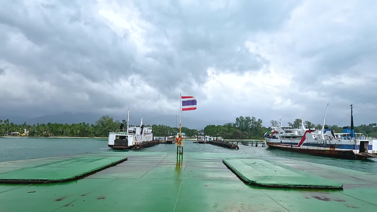 A ferry approaches the dock in Koh Samui, Thailand, under overcast skies. The Thai flag waves prominently, adding a vibrant touch