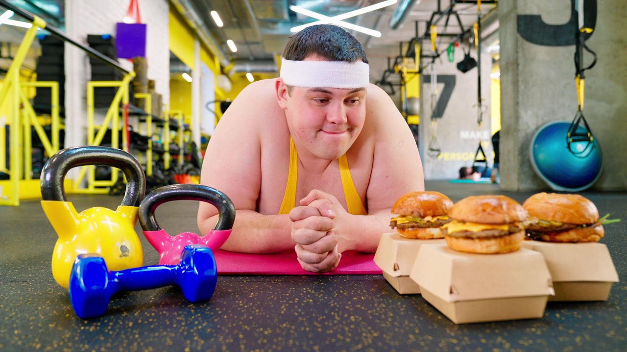 Uomo in sovrappeso su un tappetino da yoga tentato da hamburger di fast food in palestra