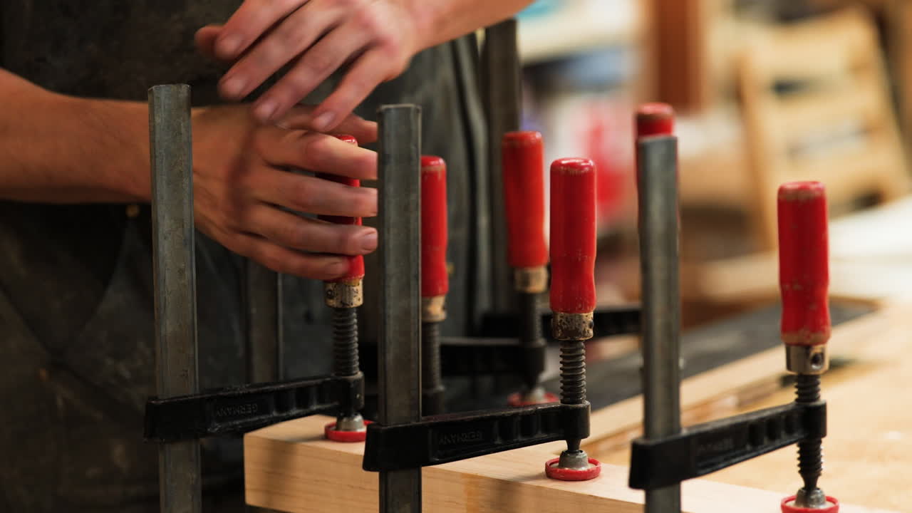 A skilled artisan carefully applies pressure with clamps to hold wooden components in place while assembling a handcrafted project in a bright workshop.