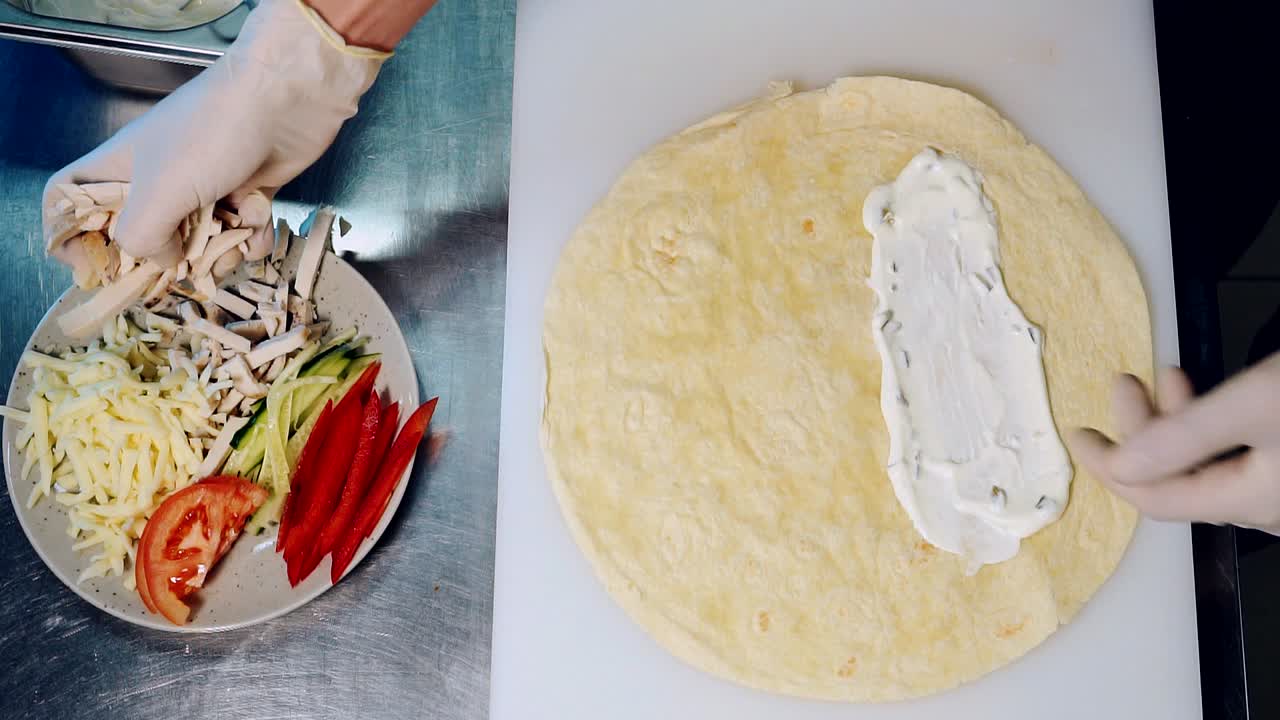 Cooking process in fast food restaurant. Hands of a chef spreading mayonnaise with a spoon and put chicken meat on pita. Top view