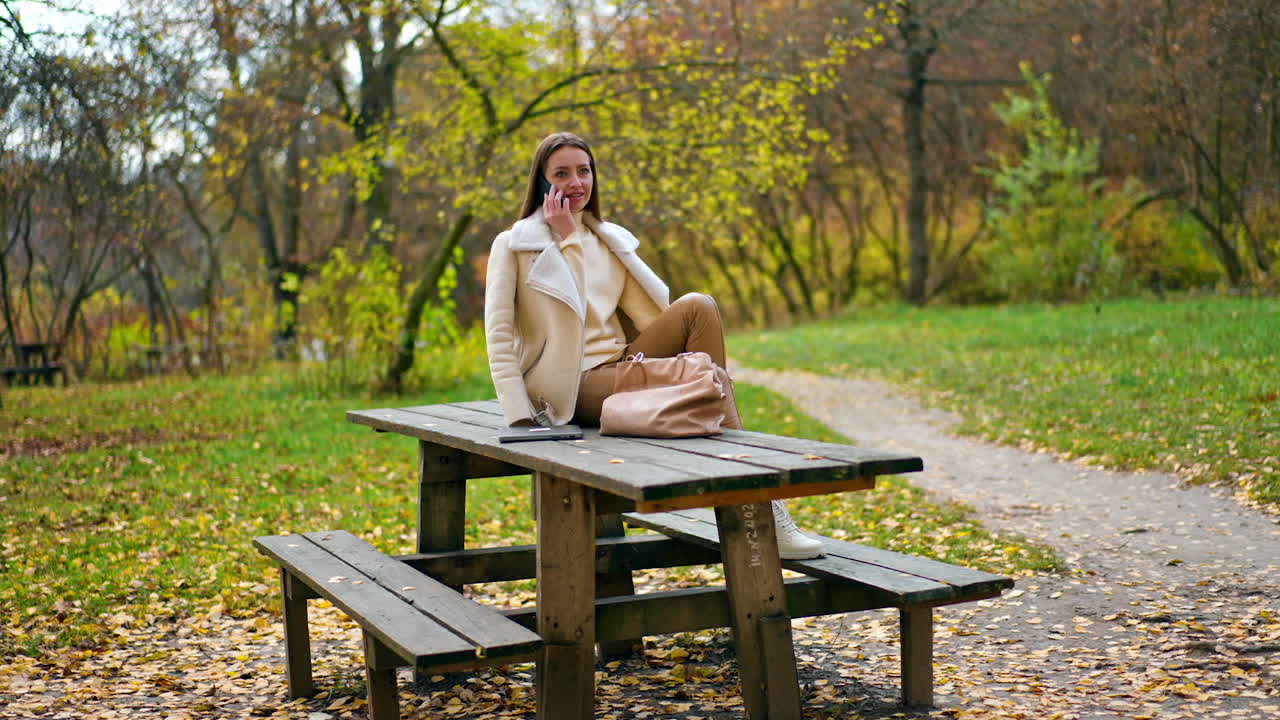 Mid-aged Caucasian lady in beige clothes sits on the wooden table in the park. Woman looking at the phone in her hand and saying something.