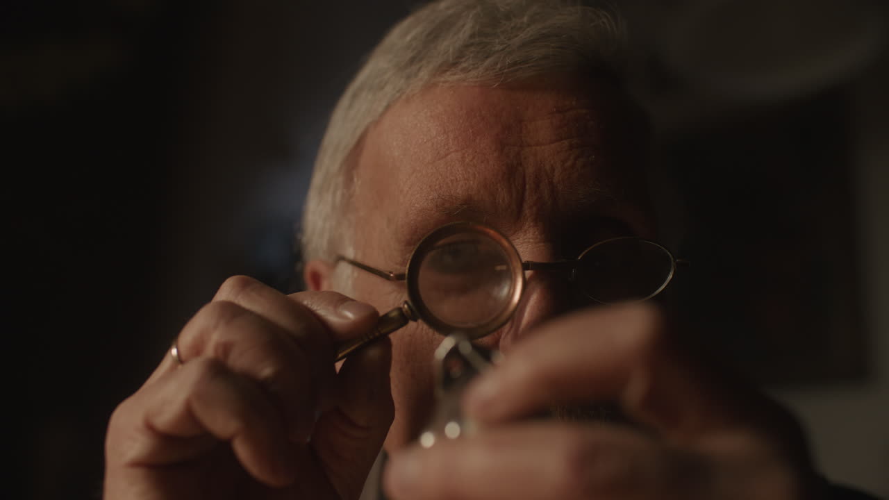 Older passionate man evaluates an old silver skull watch in his office at night