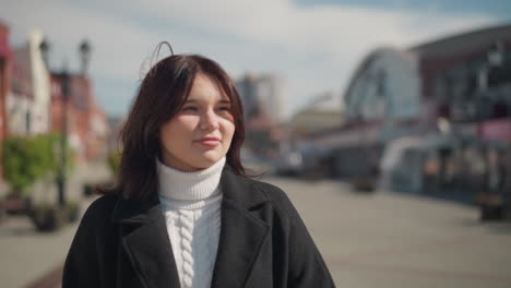 close-up of a smiling woman in winter outfit standing outdoors on a sunny day in urban setting, with blurred background of city buildings