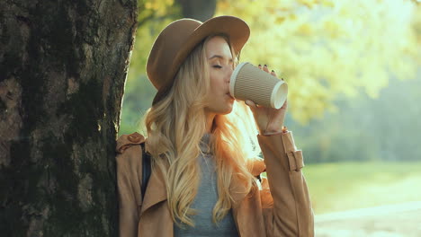 Close-up-view-of-blonde-woman-wearing-hat,-leaning-on-a-tree,-drinking-a-coffee-to-go,-and-looking-at-the-camera-with-a-smile-on-her-face-in-the-park-in-autumn