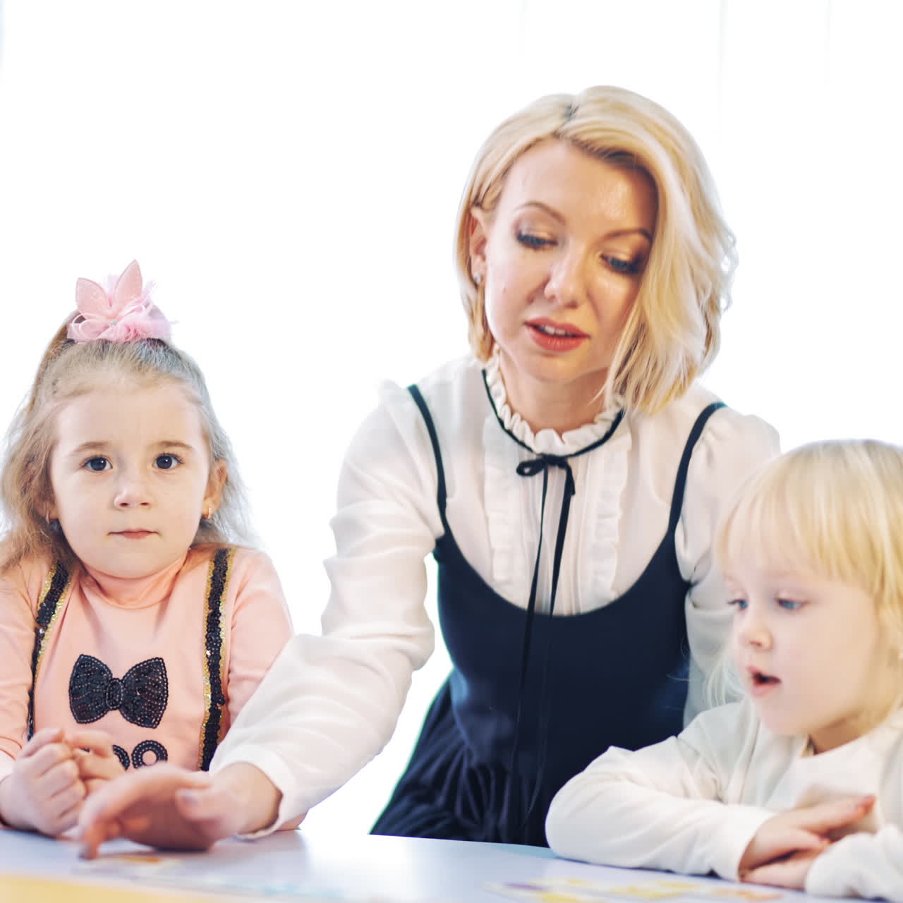 Cute preschoolers and their teacher at the table. Blonde teacher controls the work of kids and helps them to study. Woman plays with little children together indoors.
