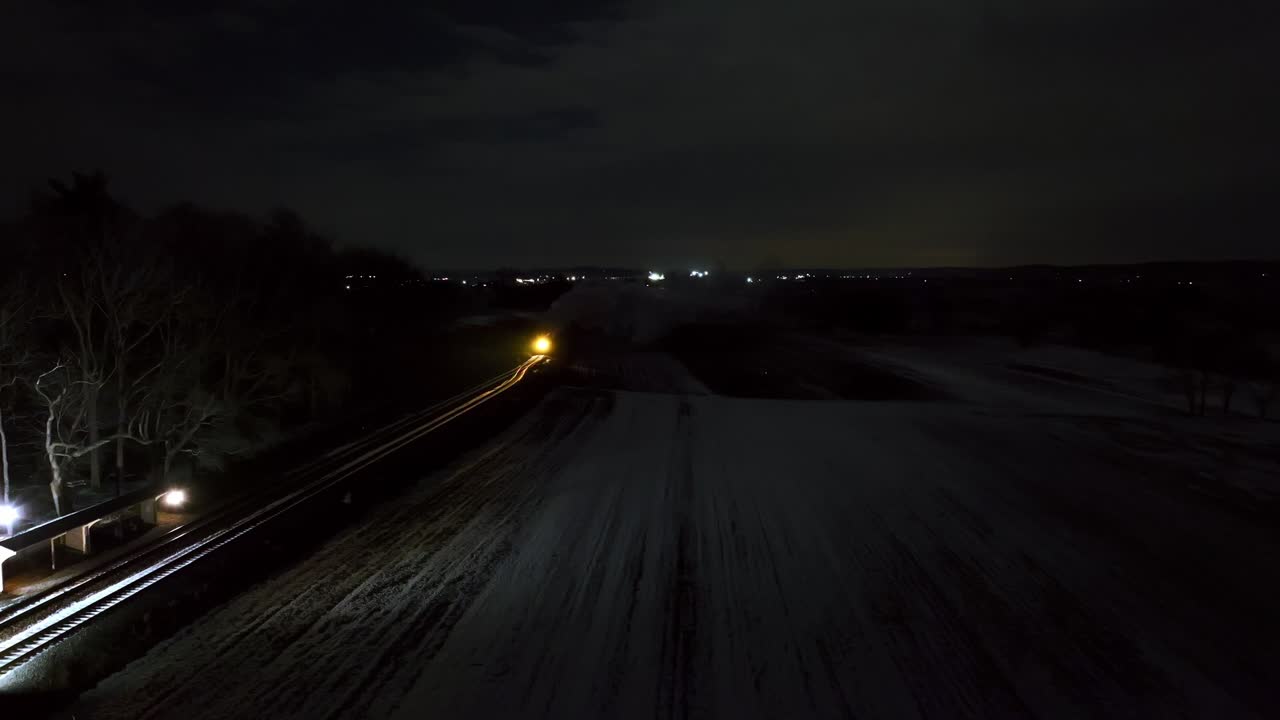 A train moves along tracks in a remote area, surrounded by fields under a dark sky. Distant lights twinkle as the train illuminates the path, creating a serene atmosphere.