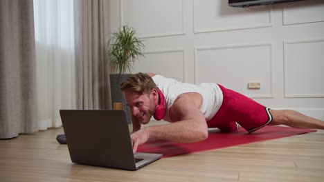 A-happy-confident-man-with-brown-hair-with-stubble-and-an-athletic-build-in-a-white-T-shirt-and-red-shorts-does-push-ups-with-one-hand-while-typing-text-on-his-laptop-with-the-other-hand-during-his-morning-exercises-in-a-modern-apartment-during-the-bright-day