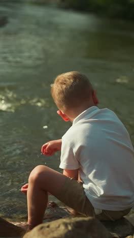 adorable boy touches water with hands on lake bank. child enjoys stunning landscape with clear water of mountain river. concept of loneliness at waterfront